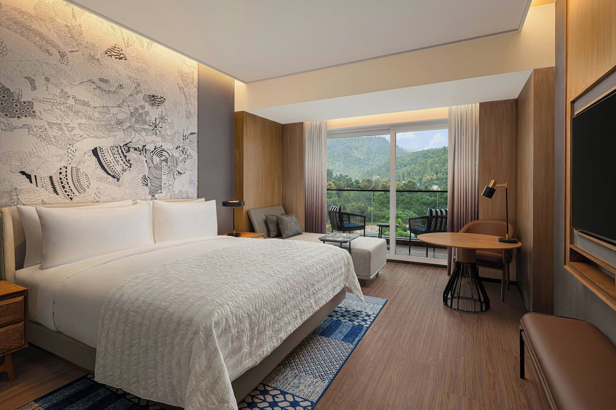 A hotel room with a large bed, a small table, a chair, a sofa, a balcony with a view of the mountains, and a wall with a detailed mural.