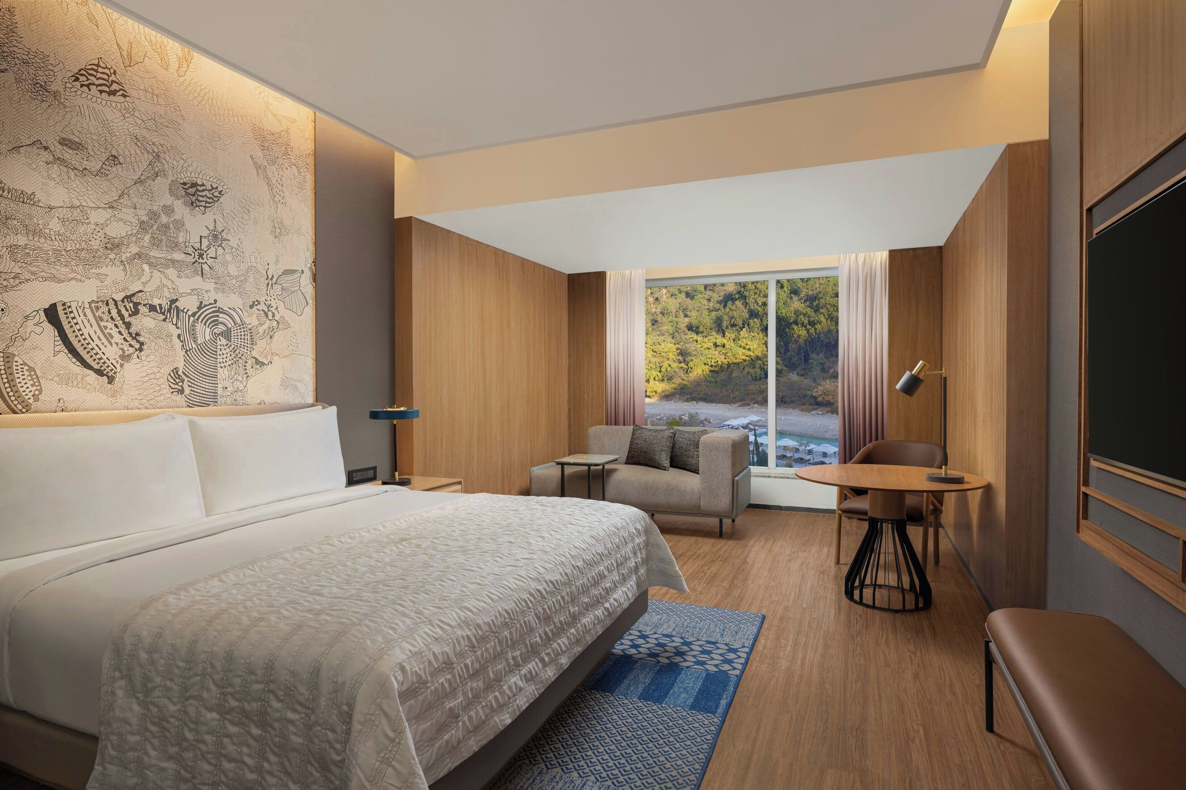 A modern hotel room with a large bed, a small table, a sofa, and a view of a forest through a large window.
