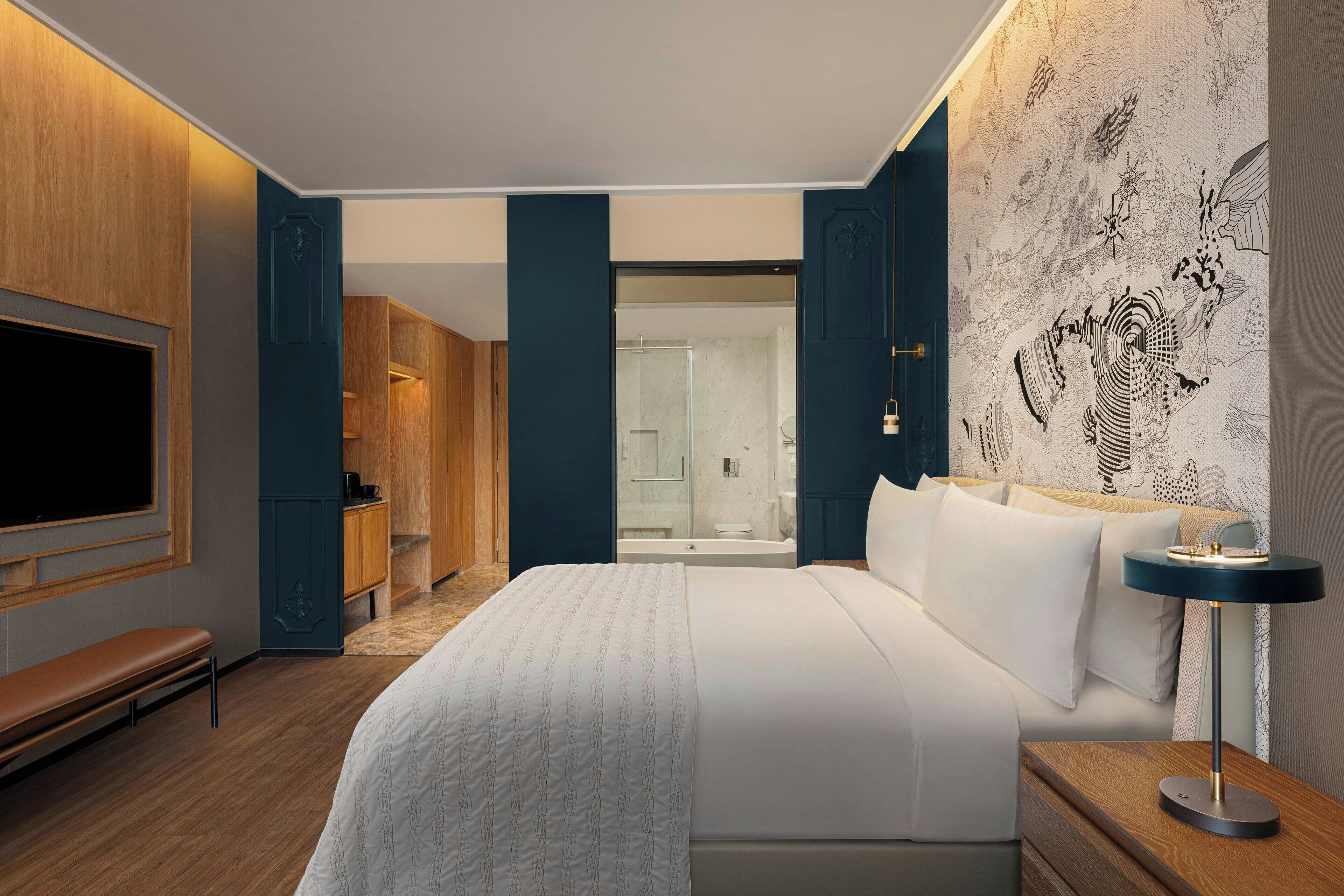 Una habitación de hotel moderna con una cama grande, un televisor montado en la pared, un banco, una mesita de noche con lámpara y una zona de ducha visible a través de una puerta de cristal.