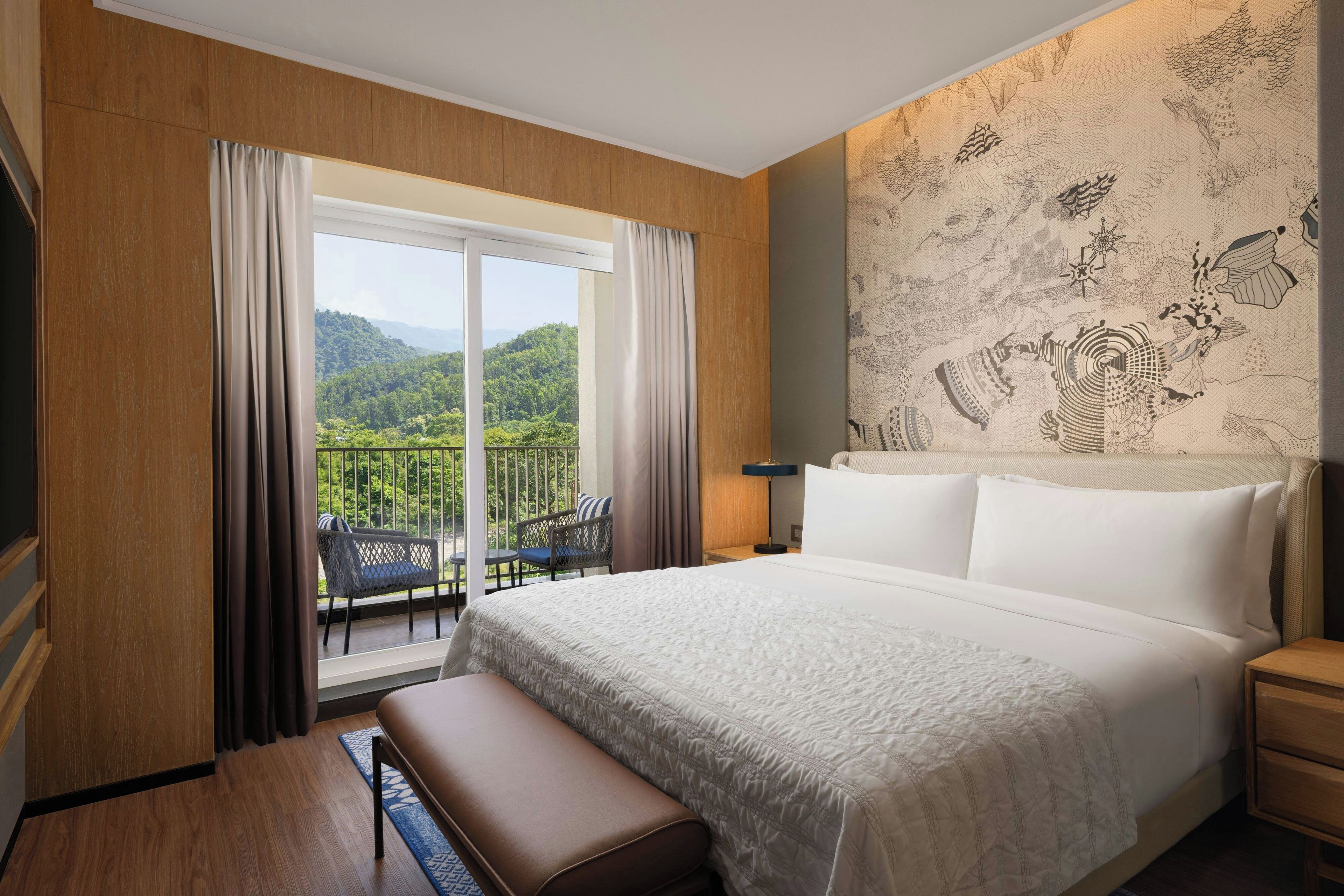 A hotel room with a large bed, a bench, a nightstand, a lamp, and a view of a mountainous landscape through a sliding door.