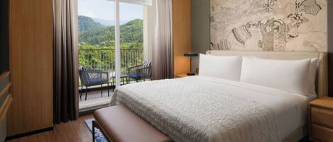 A hotel room with a large bed, a bench, a nightstand, a lamp, and a view of a mountainous landscape through a sliding door.