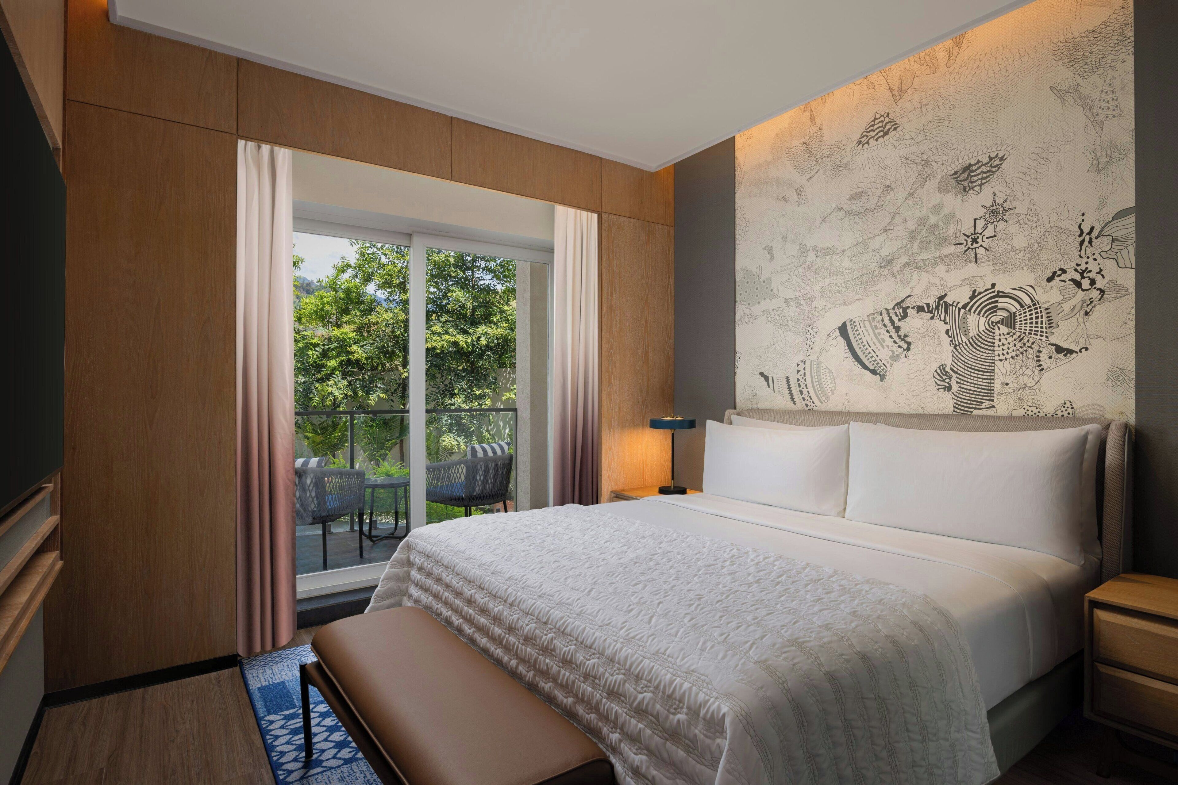 Une chambre d’hôtel dotée d’un grand lit, d’une banquette, d’une table de chevet, d’une télévision et d’un balcon aménagé avec du mobilier d’extérieur.