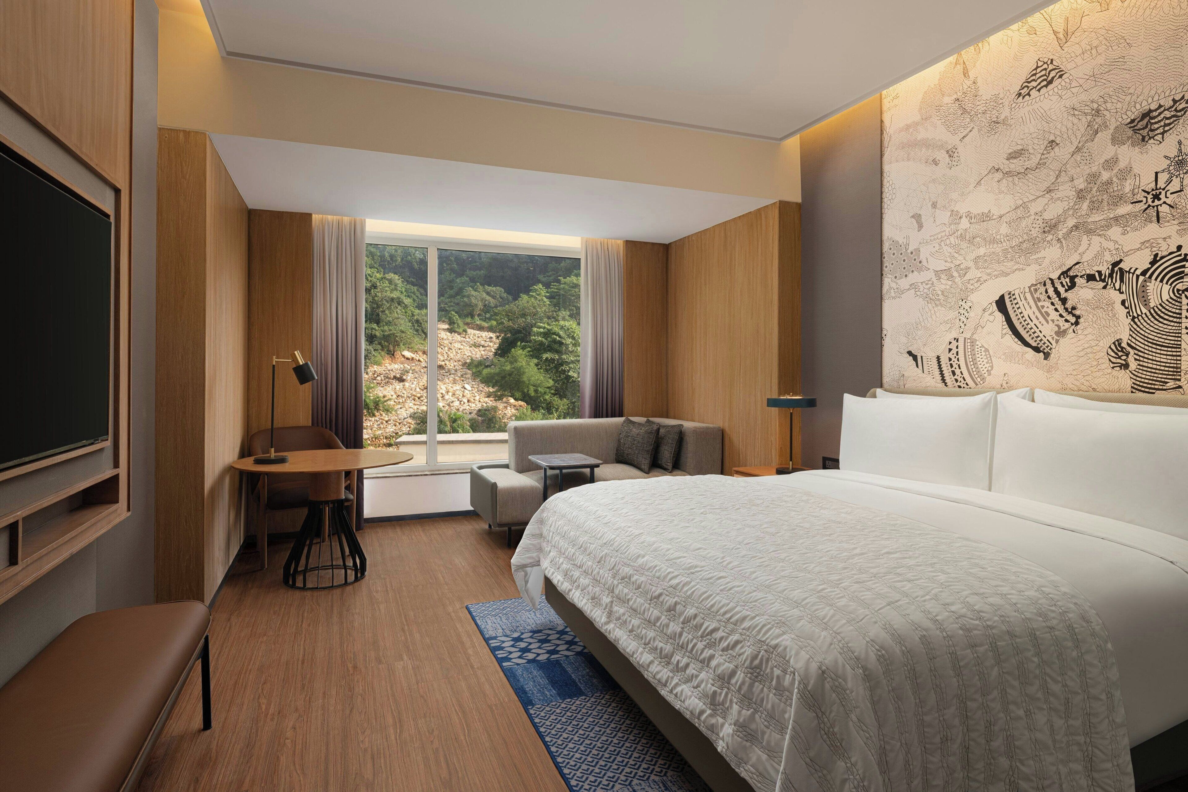 A modern hotel room with a large bed, a desk, a sofa, and a view of a mountainous landscape.