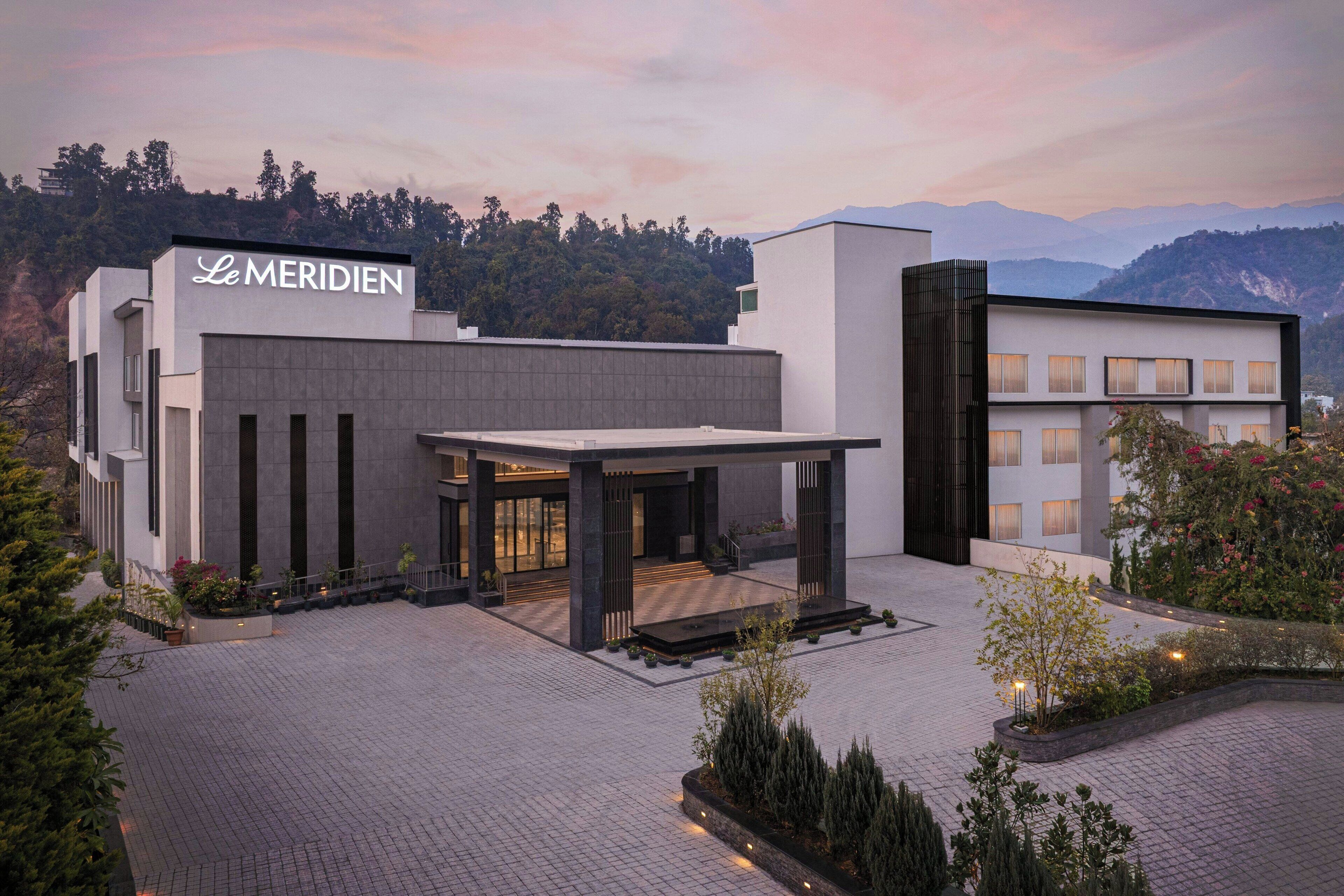 A modern hotel with a prominent 'Le MERIDIEN' sign, featuring a spacious entrance, a covered outdoor area, and a view of mountains in the background.