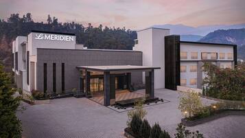A modern hotel with a prominent 'Le MERIDIEN' sign, featuring a spacious entrance, a covered outdoor area, and a view of mountains in the background.