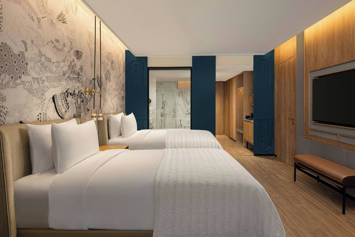 A modern hotel room with two beds, a large TV, and a bathroom visible through an open door.