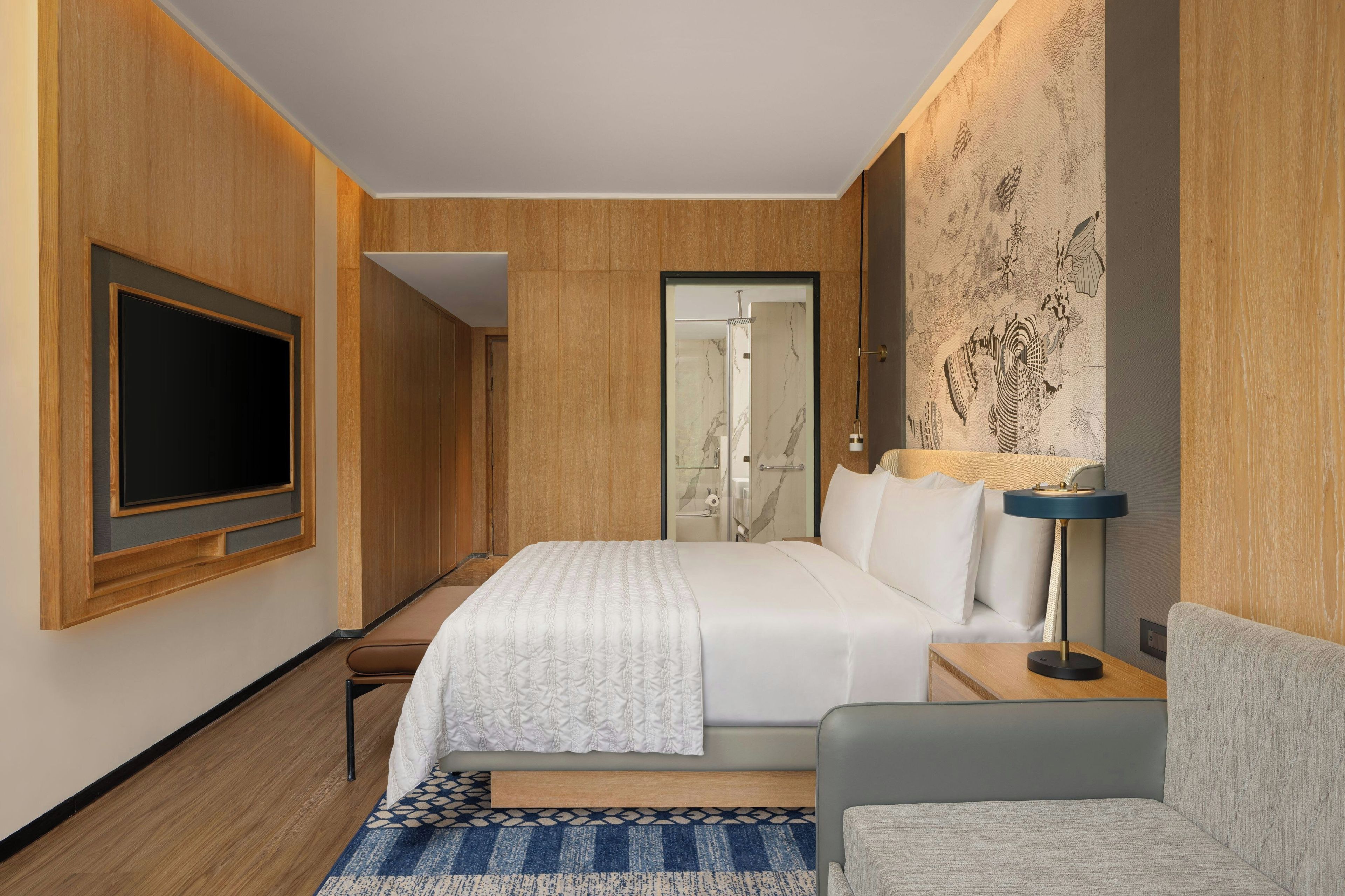 A hotel room with a bed, a television, a bedside table with a lamp, a chair, and a patterned wall.