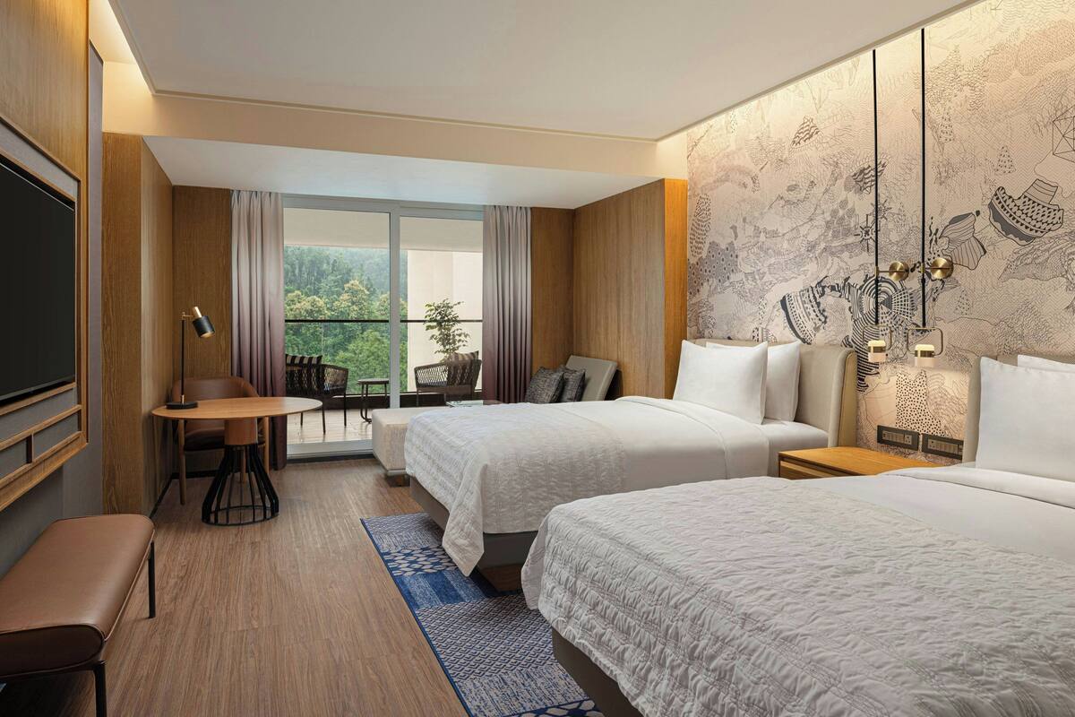 A hotel room with two beds, a desk, a chair, a TV, and a balcony with a view of greenery.