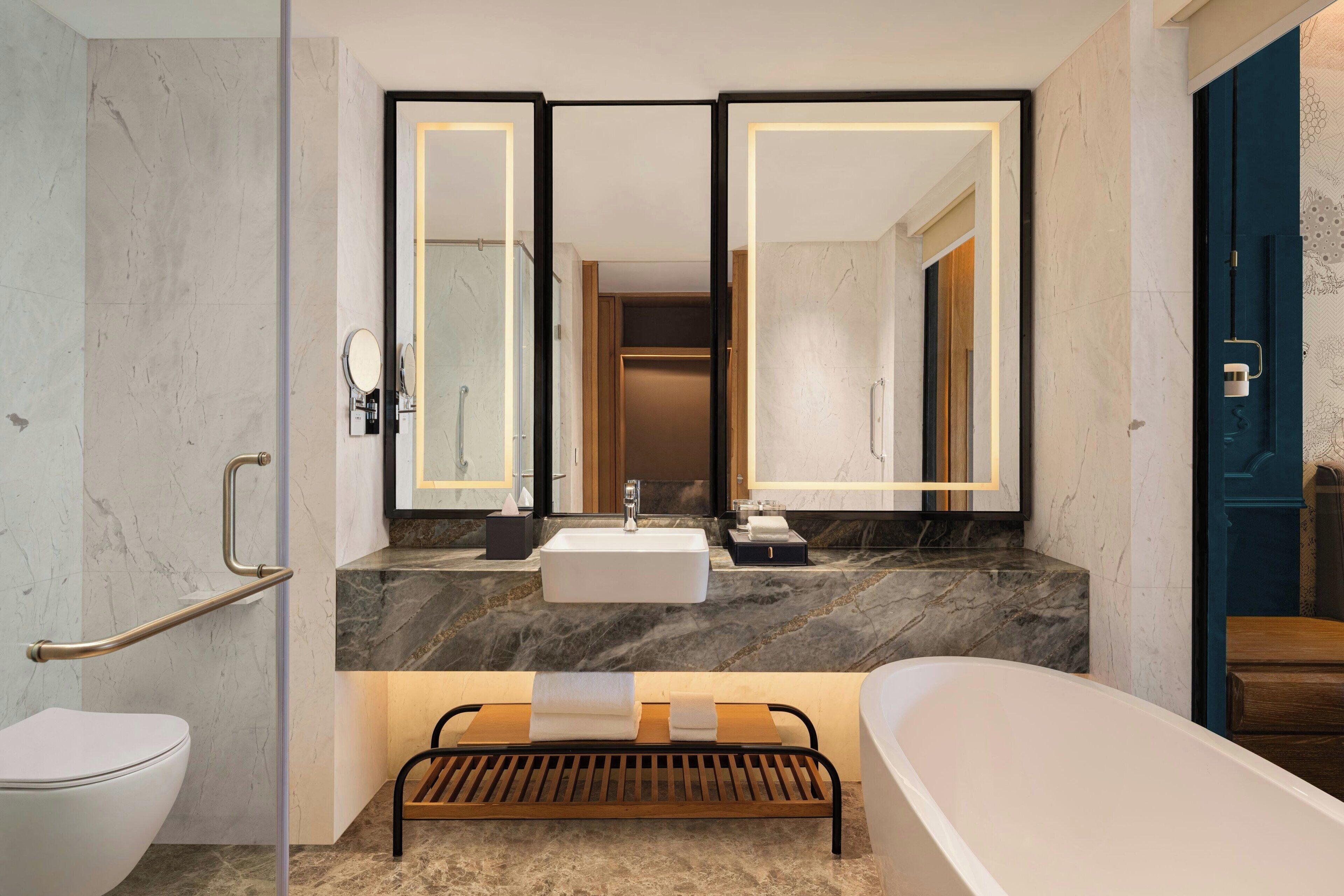 A modern bathroom with a white toilet, a sink with a mirror above it, a bathtub, and a wooden bench.