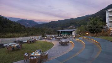An outdoor dining area with tables and chairs, surrounded by a curved pathway and a view of mountains.