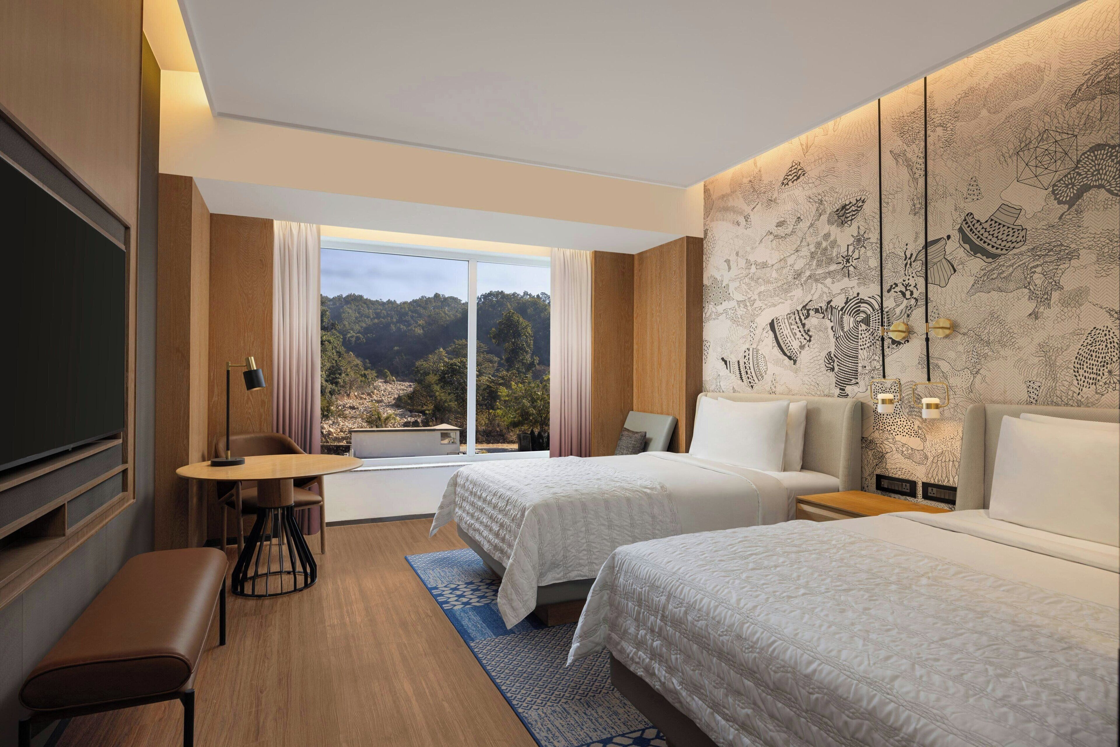 Habitación de hotel con dos camas, un escritorio, un banco, un televisor y un ventanal con vistas a un bosque.