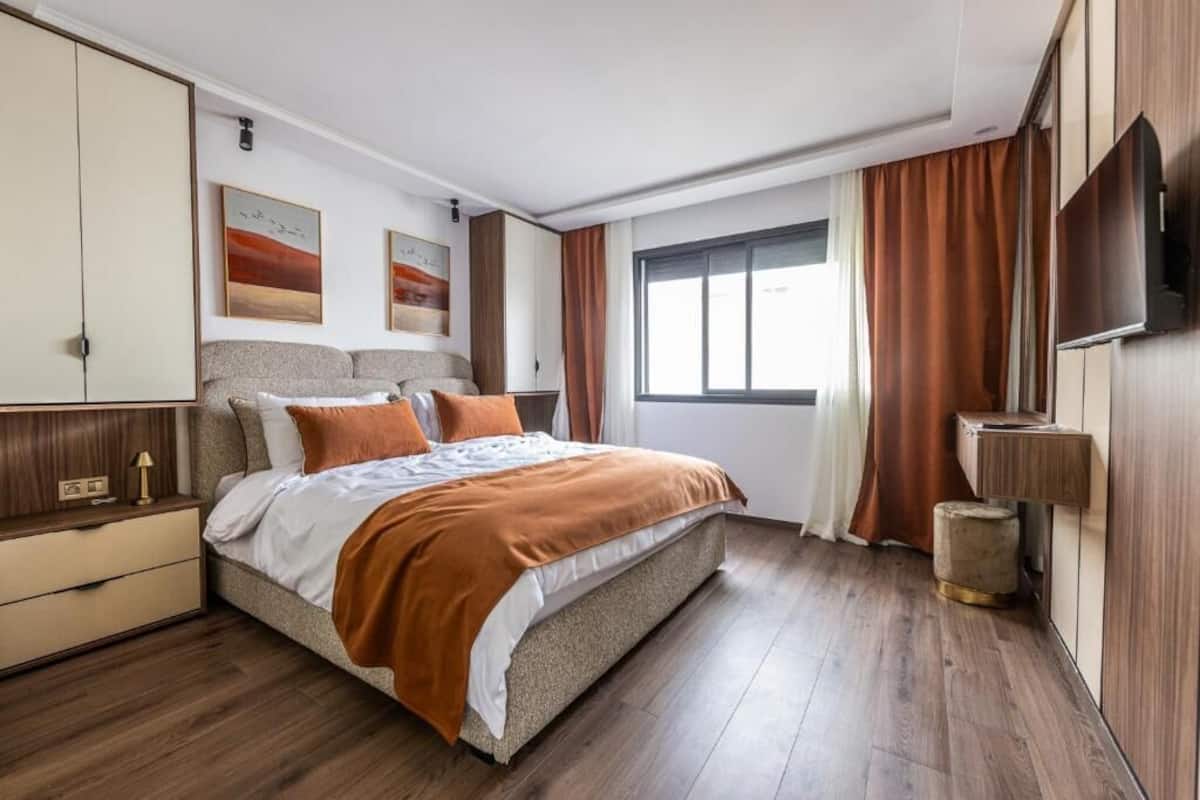 A modern bedroom with a large bed, wooden flooring, a wardrobe, a TV, and artwork on the walls.