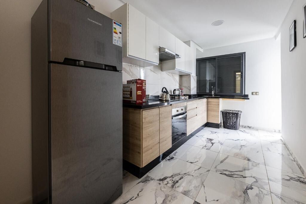 A modern kitchen with a stainless steel refrigerator, wooden cabinets, a black countertop, and a tiled floor.