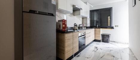 A modern kitchen with a stainless steel refrigerator, wooden cabinets, a black countertop, and a tiled floor.