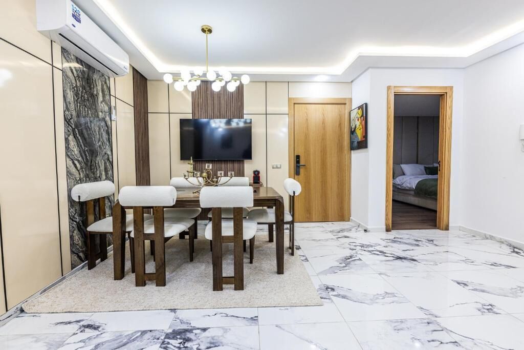 Un comedor moderno con piso de mármol, una mesa de madera con sillas blancas acolchadas, un televisor de pantalla plana montado en la pared y una lámpara en el techo.