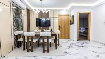 Un comedor moderno con piso de mármol, una mesa de madera con sillas blancas acolchadas, un televisor de pantalla plana montado en la pared y una lámpara en el techo.
