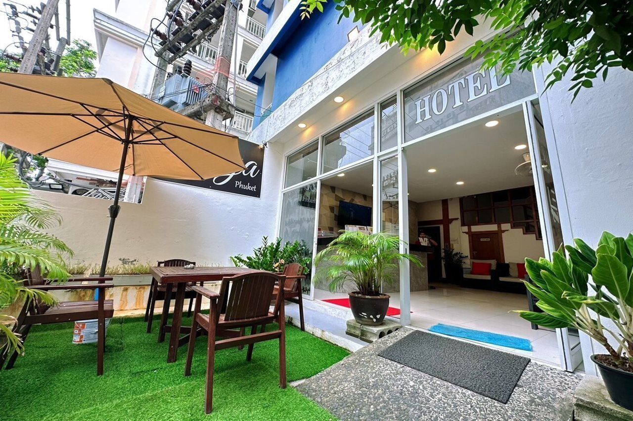 Entrada de hotel con fachada de vidrio, zona de estar al aire libre con mesa y sillas, y plantas en macetas.