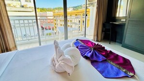A hotel room with a large window, a bed with white linens, a purple and pink decorative item, and a view of buildings and a balcony. - Hotel Baya Patong Beach (Patong)