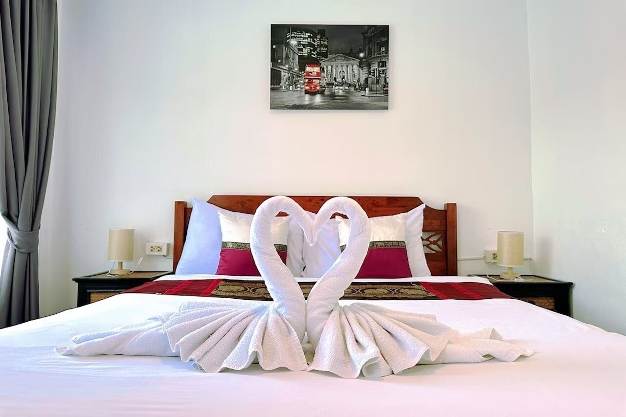 A hotel room with a neatly made bed, a heart-shaped towel arrangement, a cityscape painting on the wall, and a nightstand with a lamp.