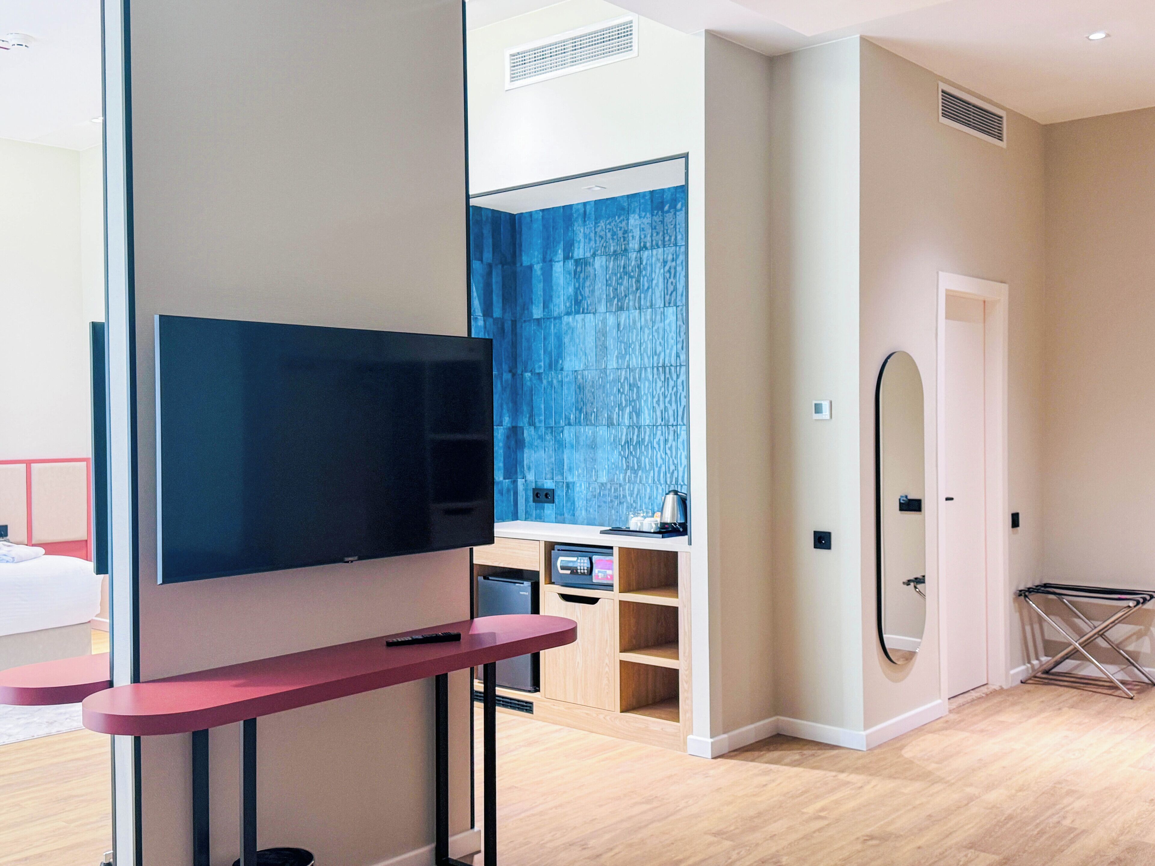 A modern hotel room with a flat-screen TV, a small kitchen area, and a bathroom with a blue-tiled wall.