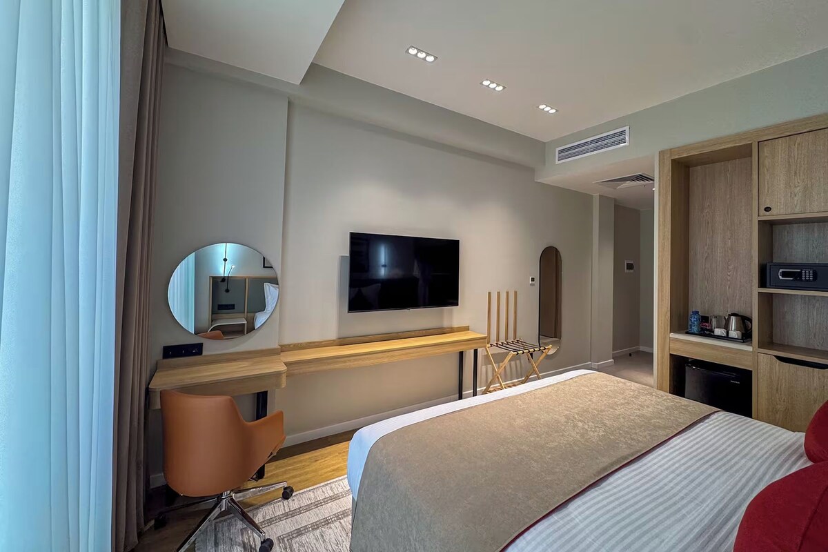A modern hotel room with a bed, a TV, a desk, a mirror, and a wardrobe.