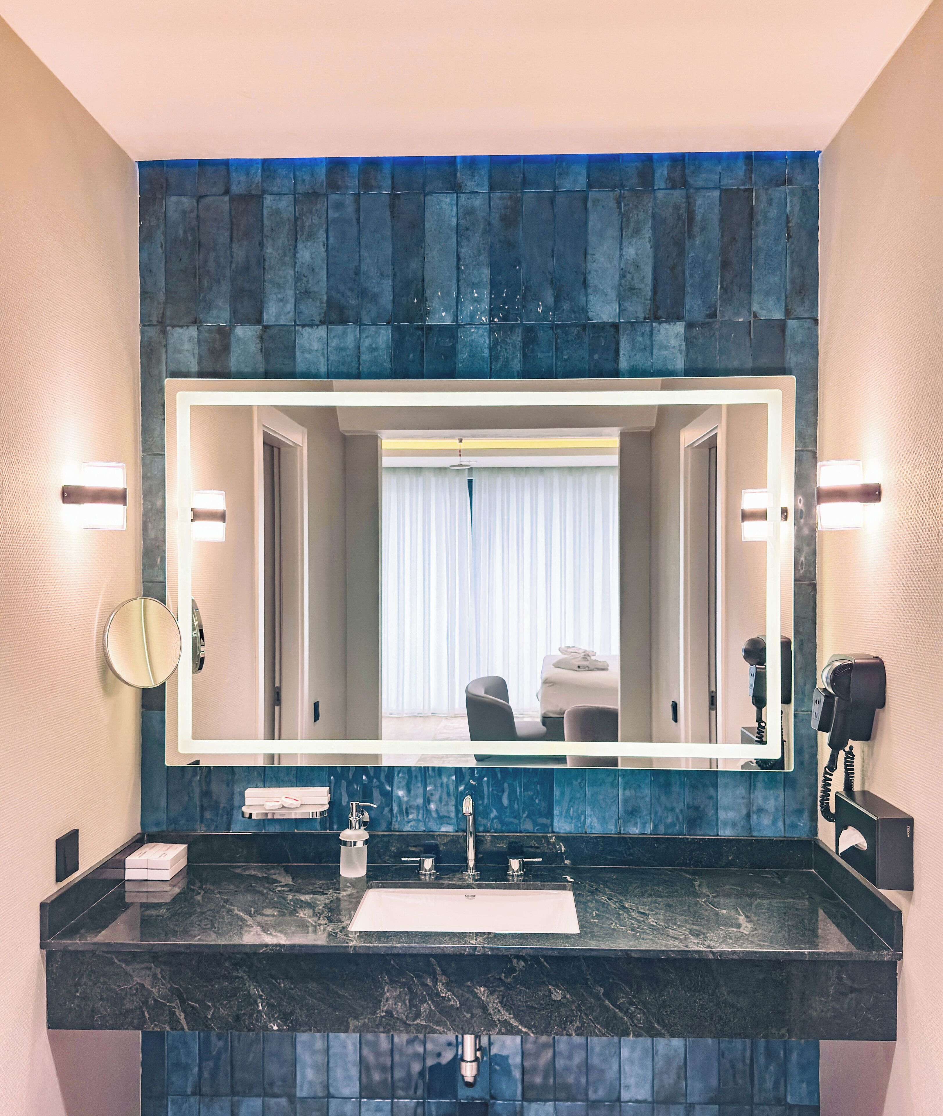 A bathroom with a large mirror, black marble countertop, and a view of a room with a bed and window.