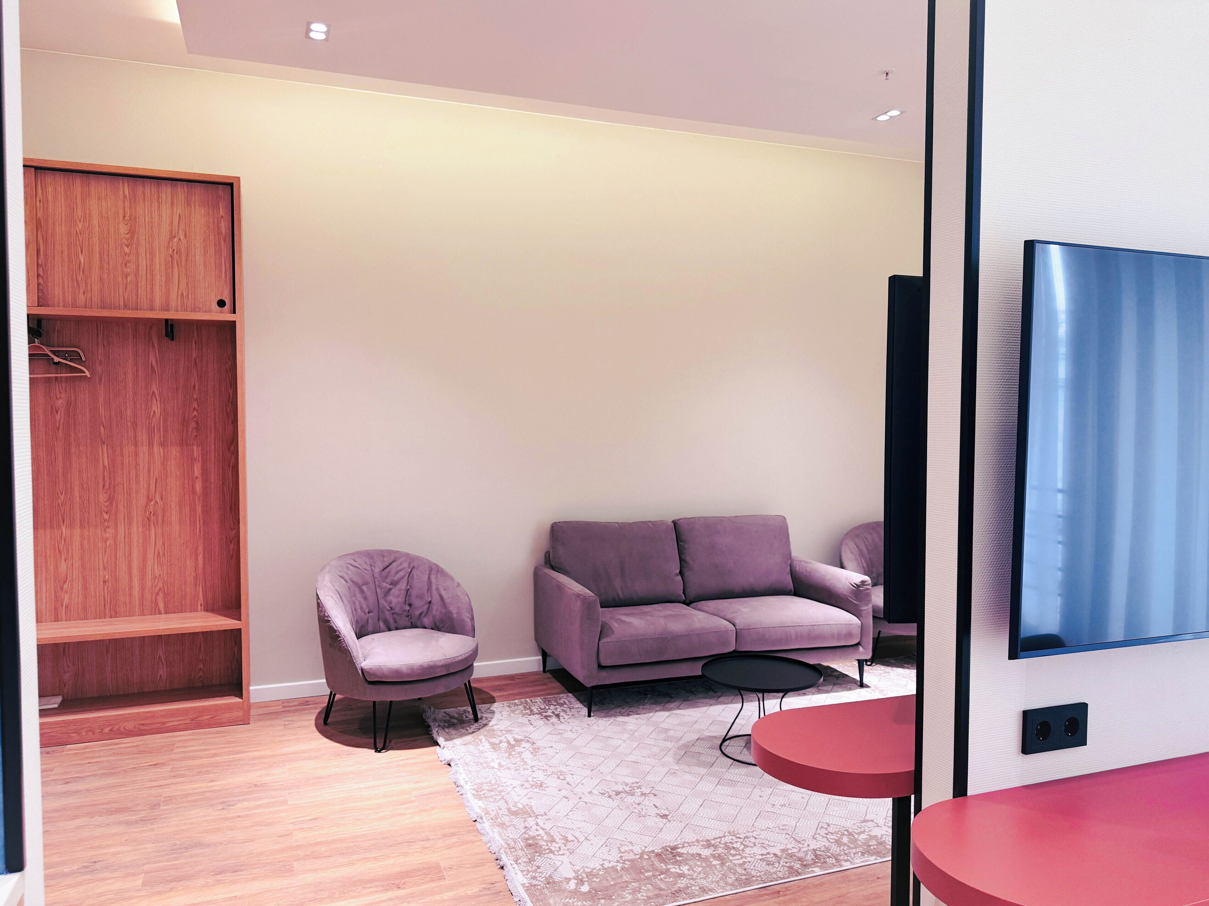 A modern room with a wooden wardrobe, a purple armchair, a sofa, a small round table, and a TV mounted on the wall.