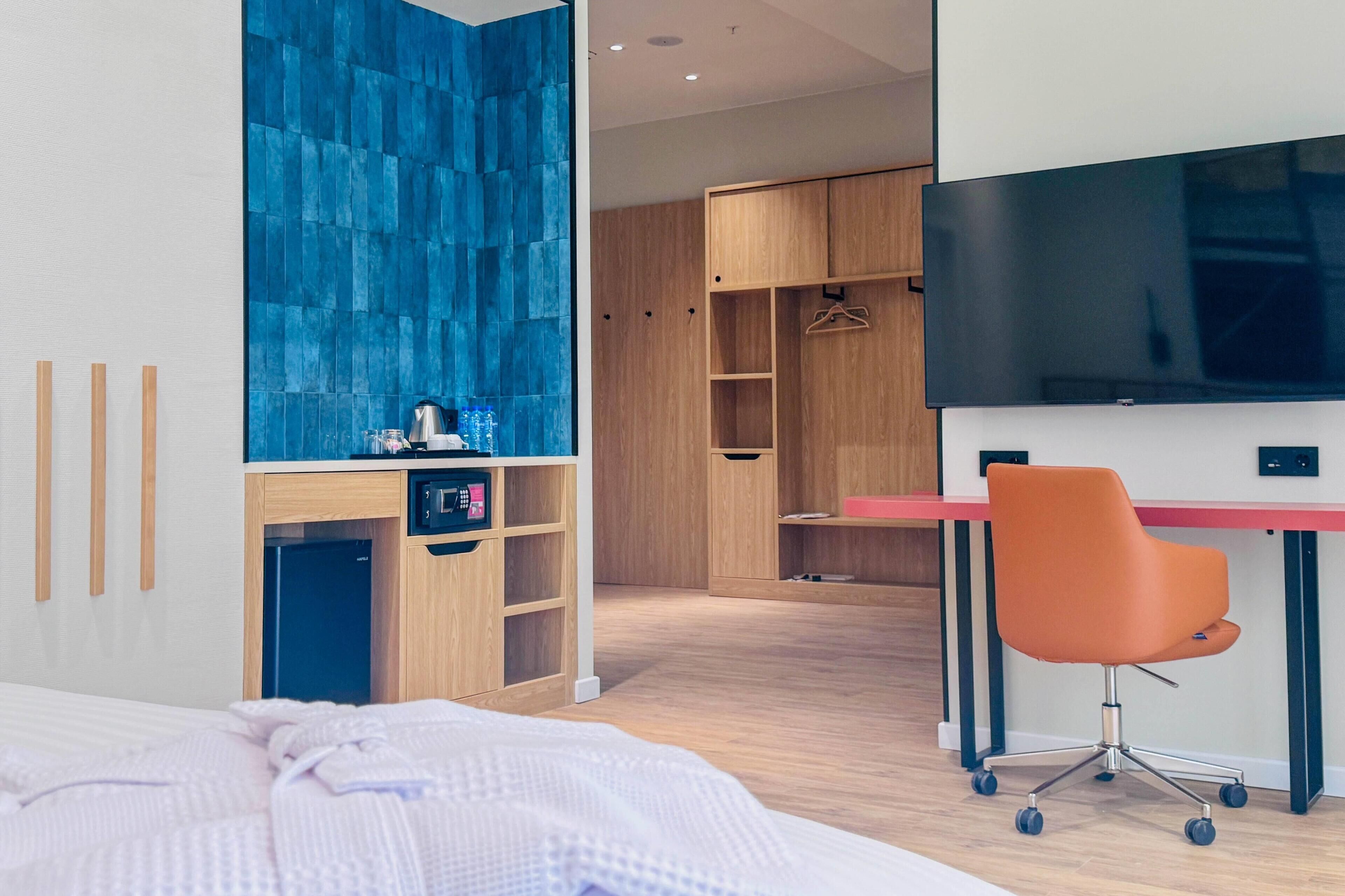 A modern hotel room with a bed, a desk, a chair, a wardrobe, and a TV.
