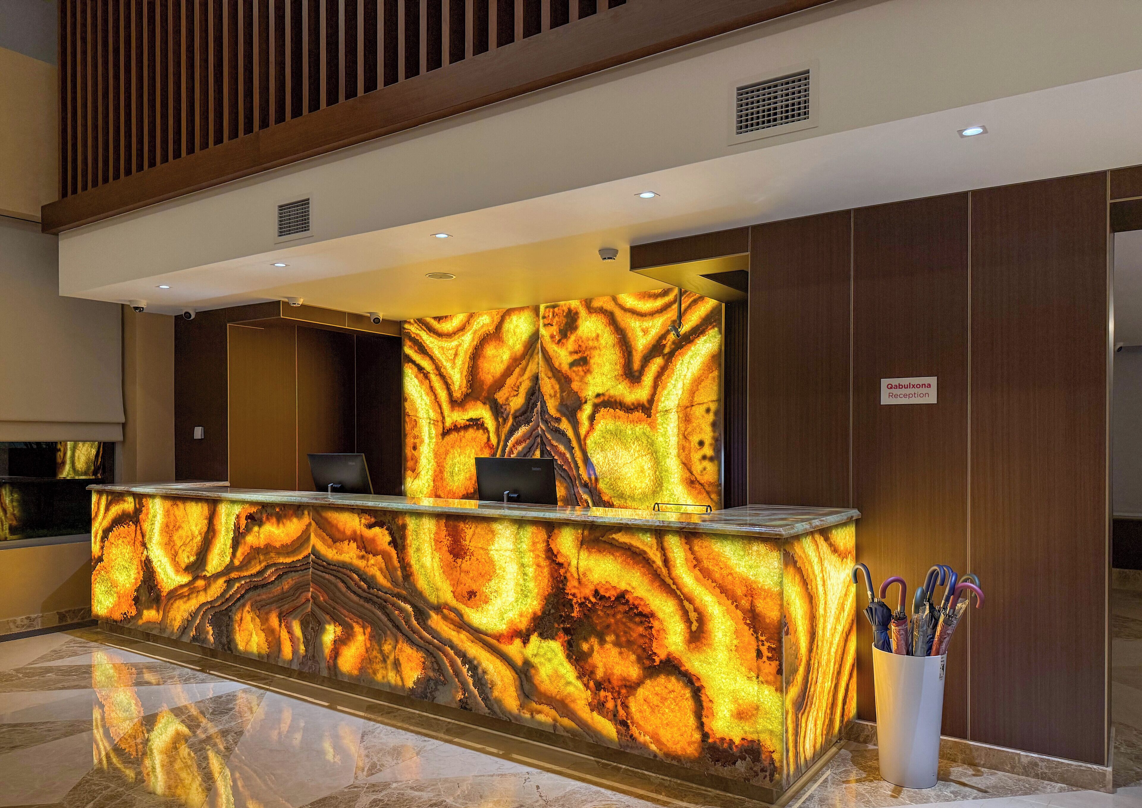 A reception desk with a marble-like design, two computer monitors, a sign for 'Gabulxona Reception', and a white umbrella holder with umbrellas.