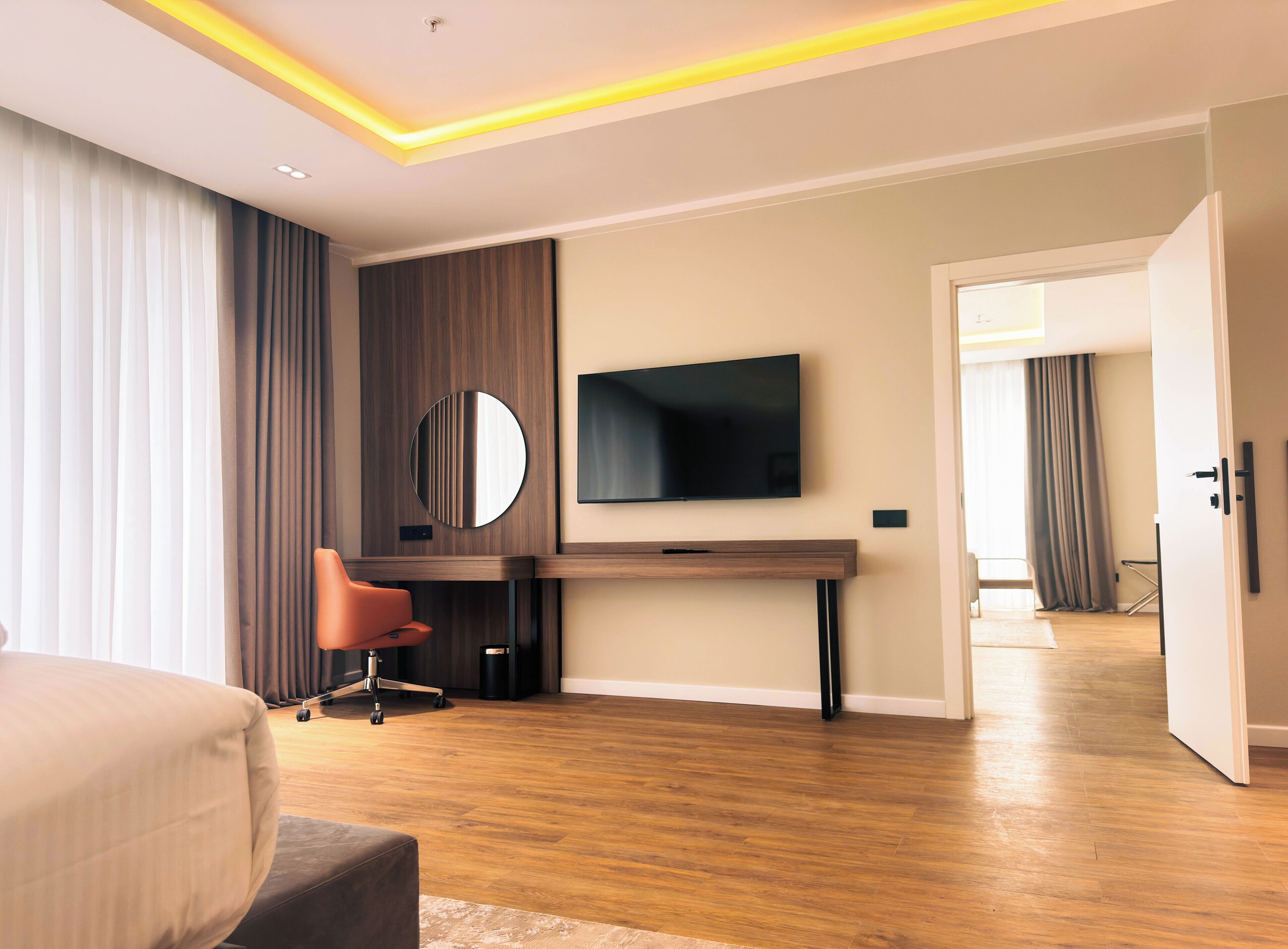 A modern hotel room with a wooden floor, a large bed, a desk with a chair, a TV mounted on the wall, and a door leading to another room.