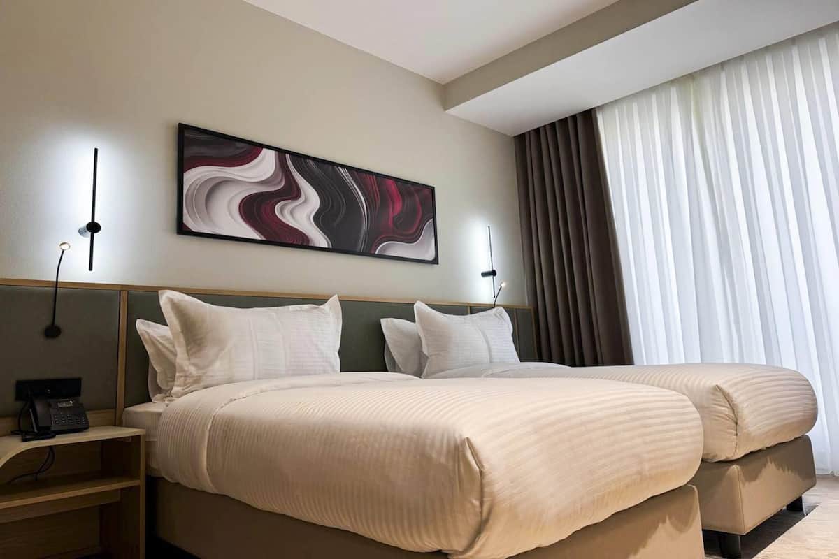 A hotel room with two beds, a modern abstract painting on the wall, a bedside table with a phone, and a large window with curtains.