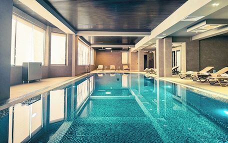 A modern indoor swimming pool with clear water, sunlit by large windows, and equipped with sun loungers along the sides.