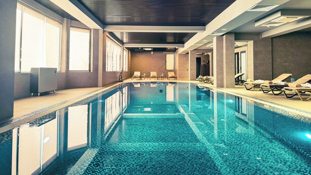 Une piscine intérieure moderne, dotée d’une eau cristalline, baignée de lumière grâce à de grandes fenêtres, et aménagée avec des chaises longues le long des bords.