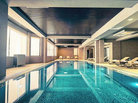 A modern indoor swimming pool with clear water, sunlit by large windows, and equipped with sun loungers along the sides.