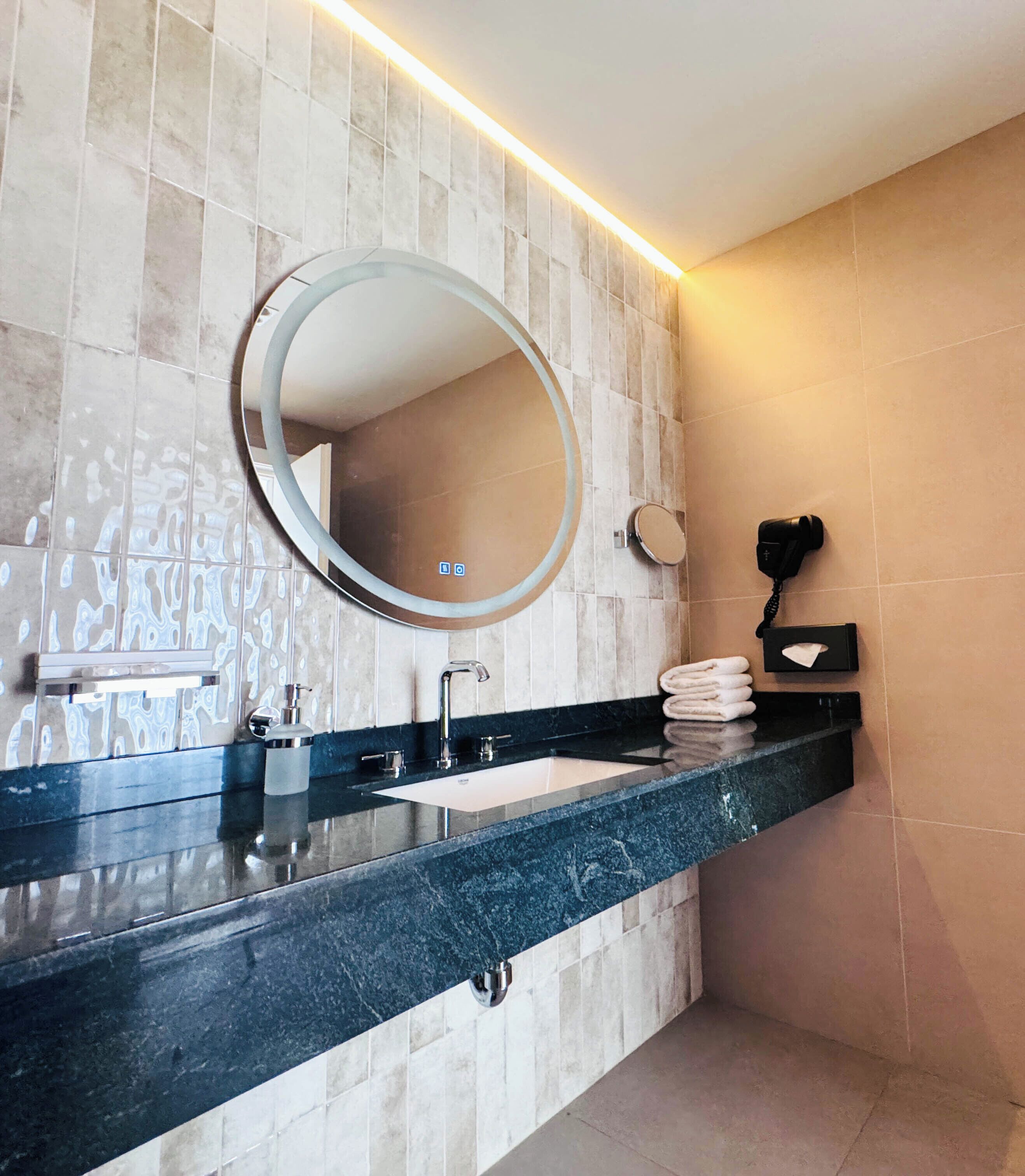 A modern bathroom with a marble vanity, a round mirror, a sink, and a hairdryer mounted on the wall.