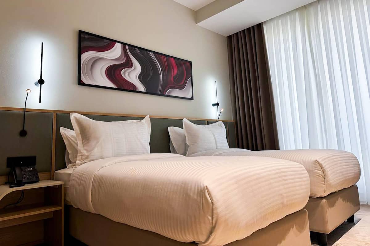 A hotel room with two beds, a painting on the wall, a telephone on the nightstand, and a window with curtains.