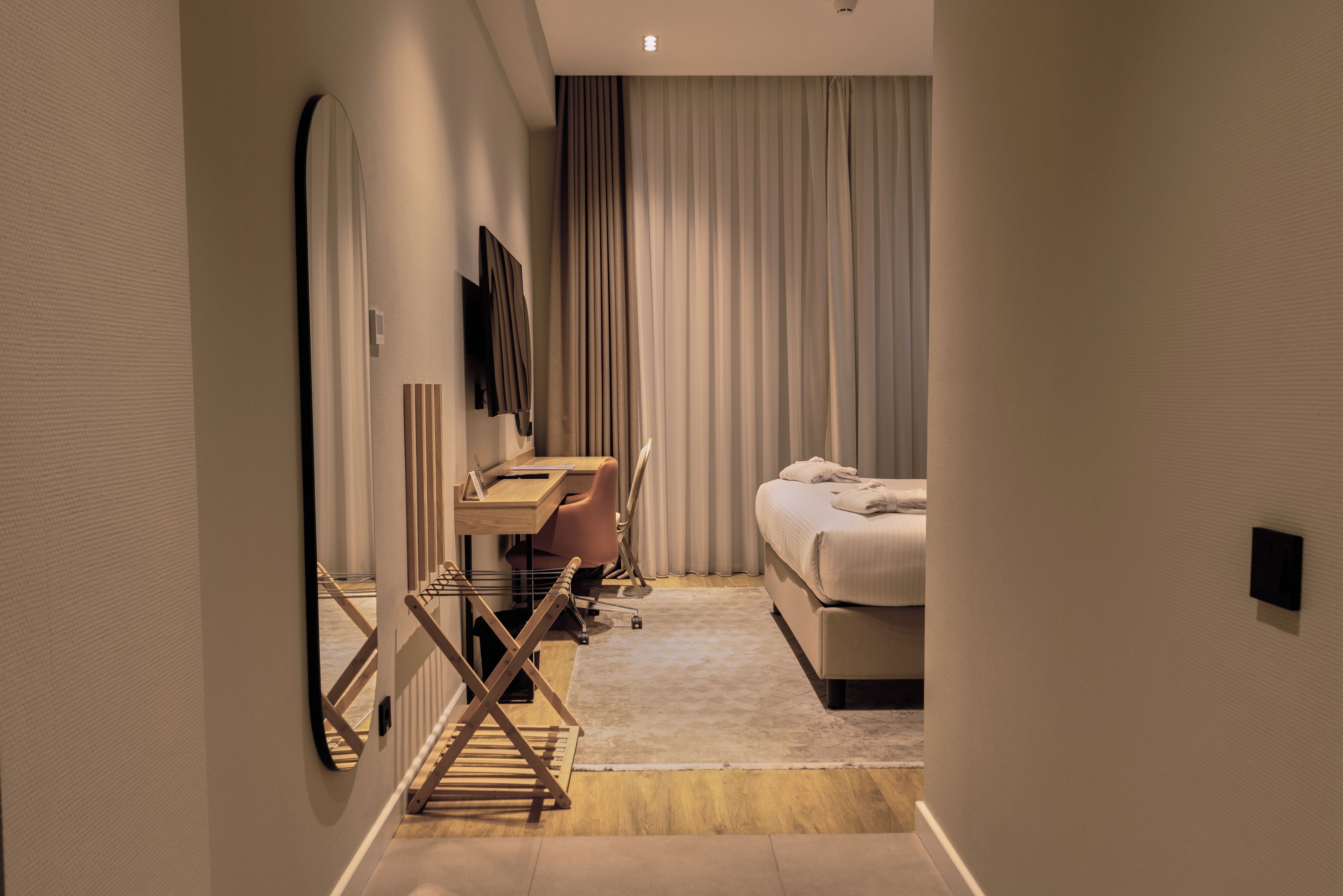 A hotel room with a bed, a desk with a chair, a television mounted on the wall, and a window with curtains.