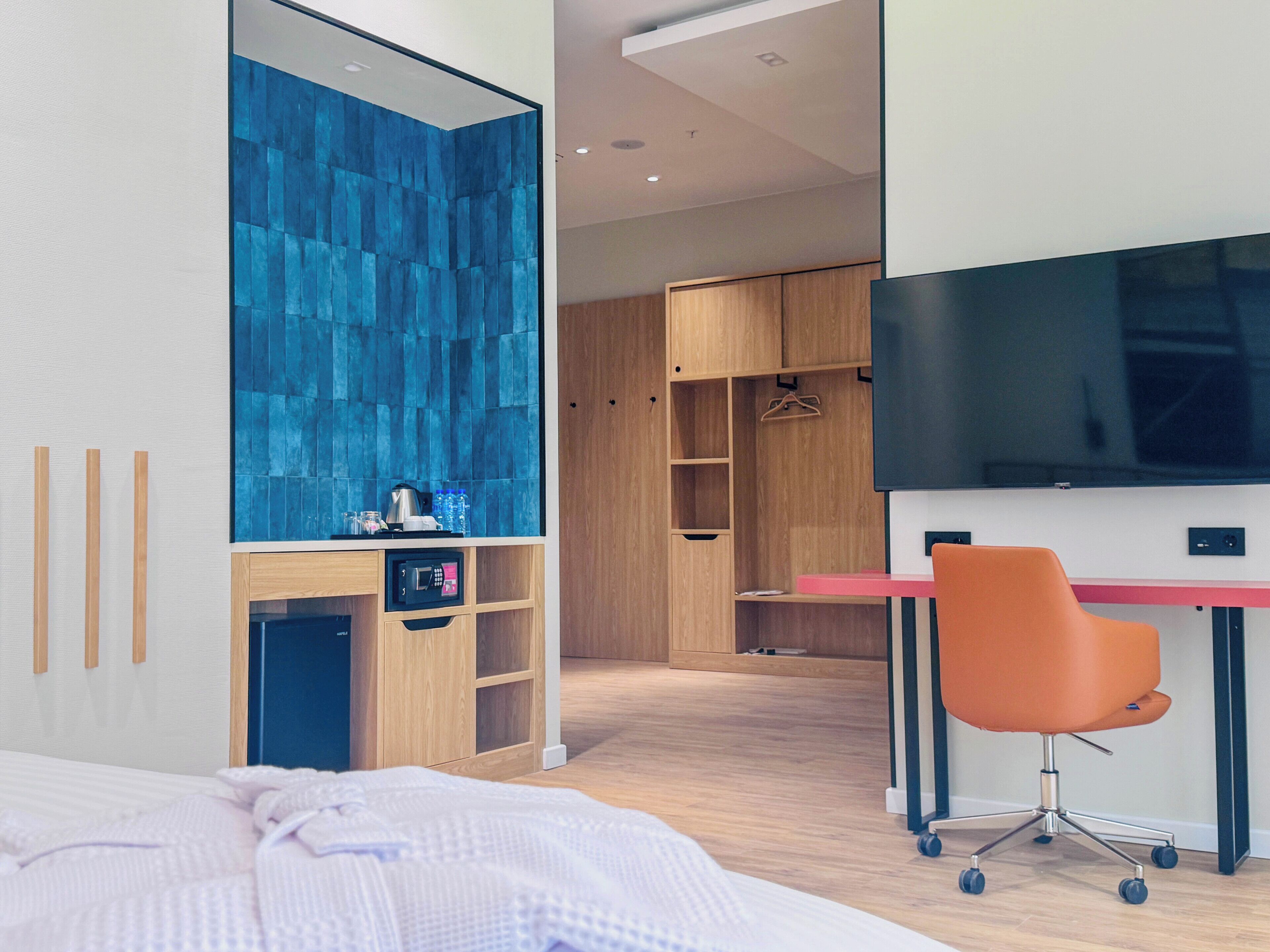 Quarto de hotel moderno com cama, escrivaninha com cadeira laranja, televisão, guarda-roupa e parede com azulejos azuis.