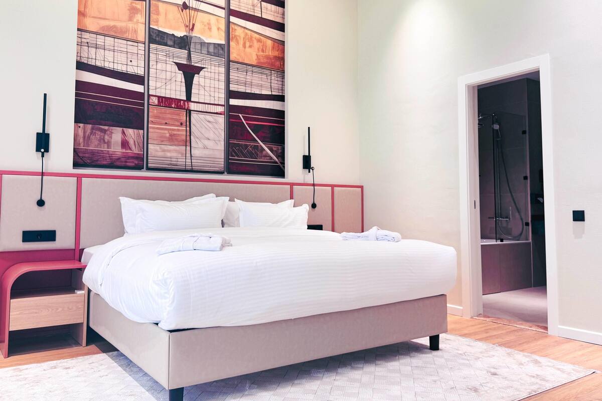 A modern bedroom with a large bed, a bedside table, a wall-mounted artwork, and a shower area visible through an open door.