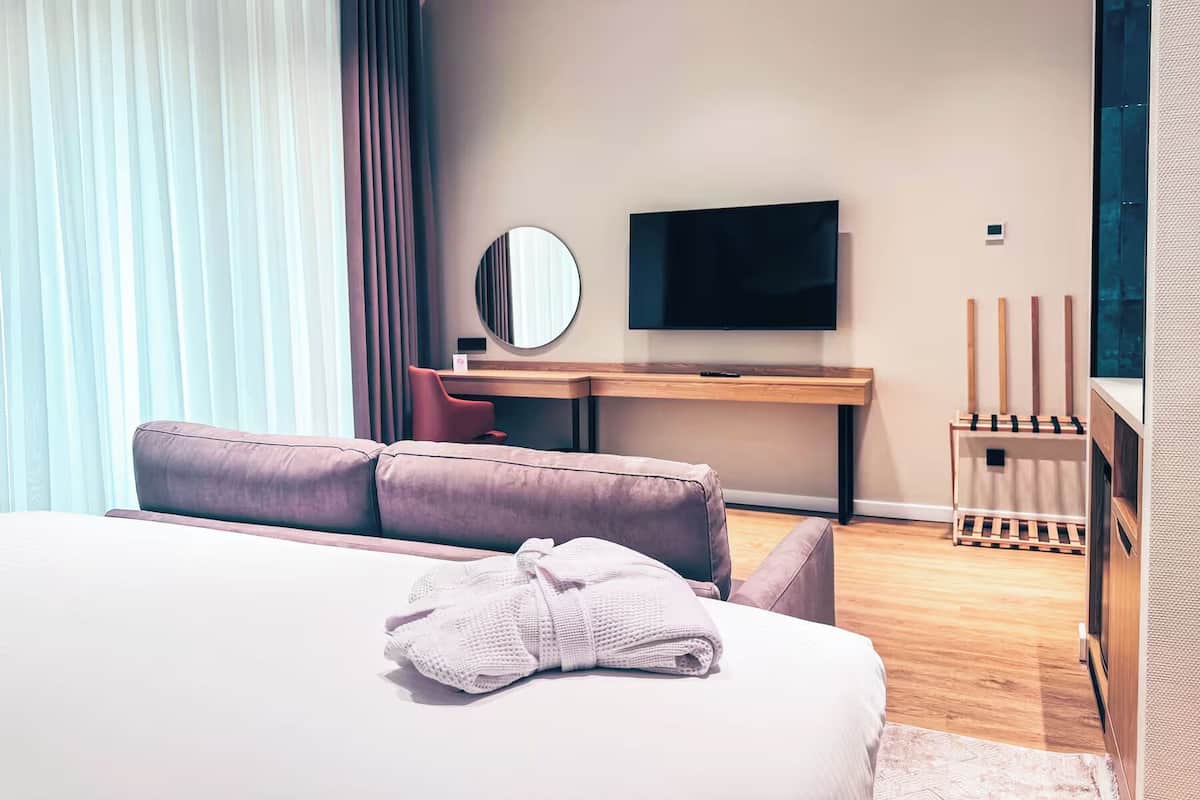 A modern hotel room with a bed, a desk, a TV, and a window with curtains.
