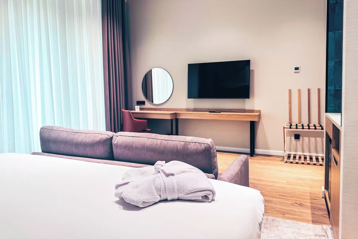 A modern hotel room with a bed, a desk, a TV, and a window with curtains.