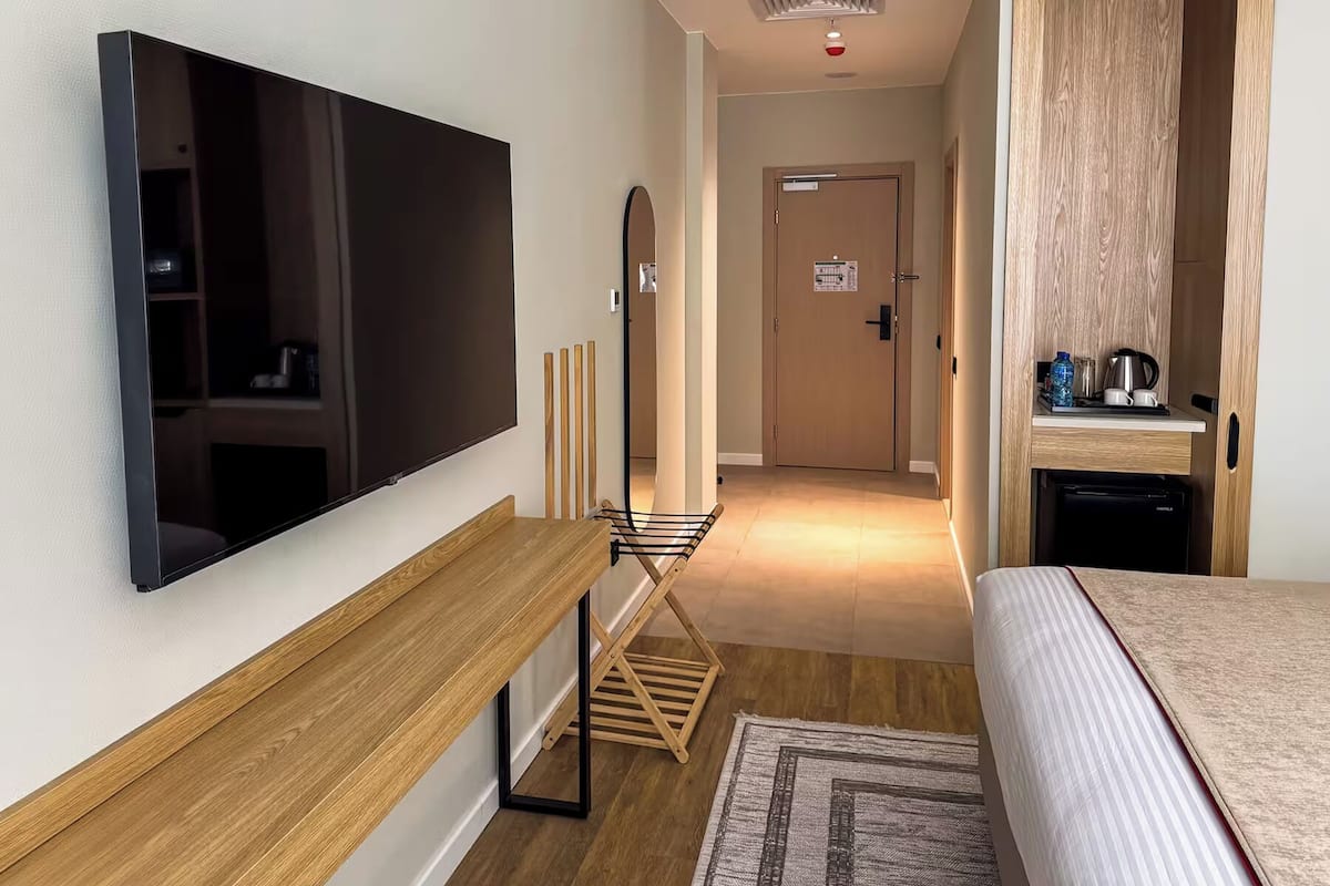 A hotel room with a bed, a TV mounted on the wall, a wooden bench, a small table with a kettle, and a door leading to another room.