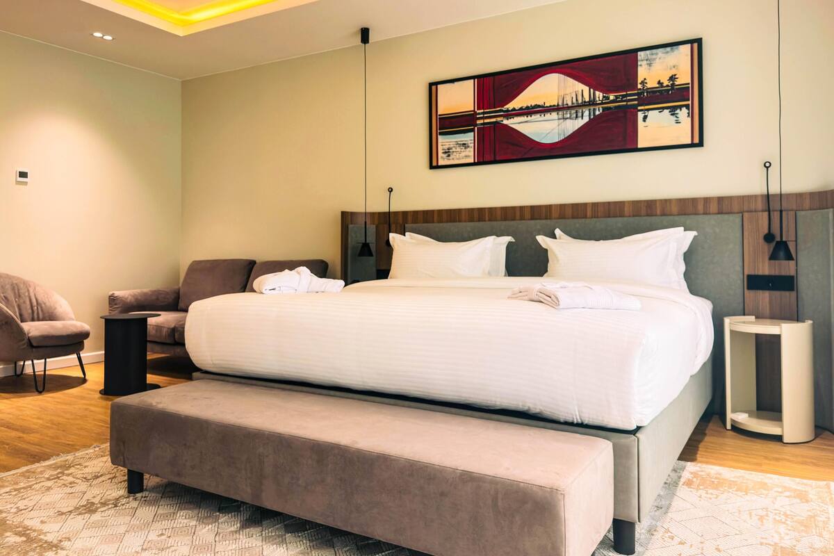 A modern hotel bedroom with a large bed, a bench, a chair, and a painting on the wall.