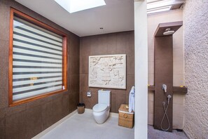 Une salle de bain équipée de toilettes, d’un bidet et d’une douche. Il y a une fenêtre avec des stores et un élément décoratif mural.
