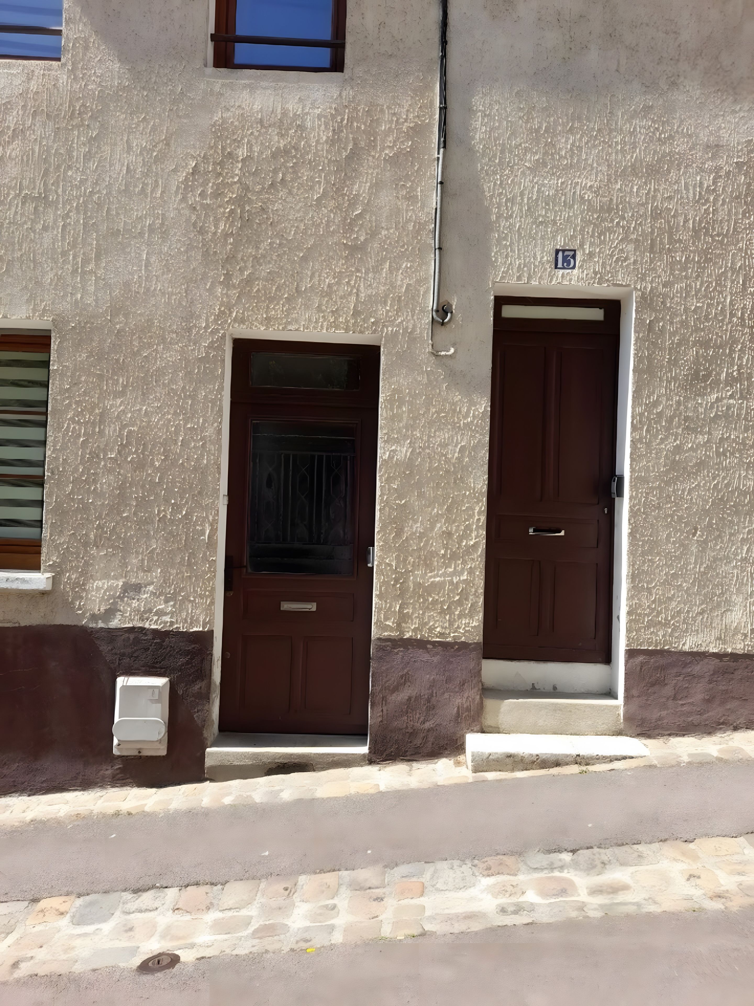 Un bâtiment avec deux portes marron, une entrée pavée de pierres et un panneau indiquant le numéro de l’immeble au-dessus de l’une des portes.