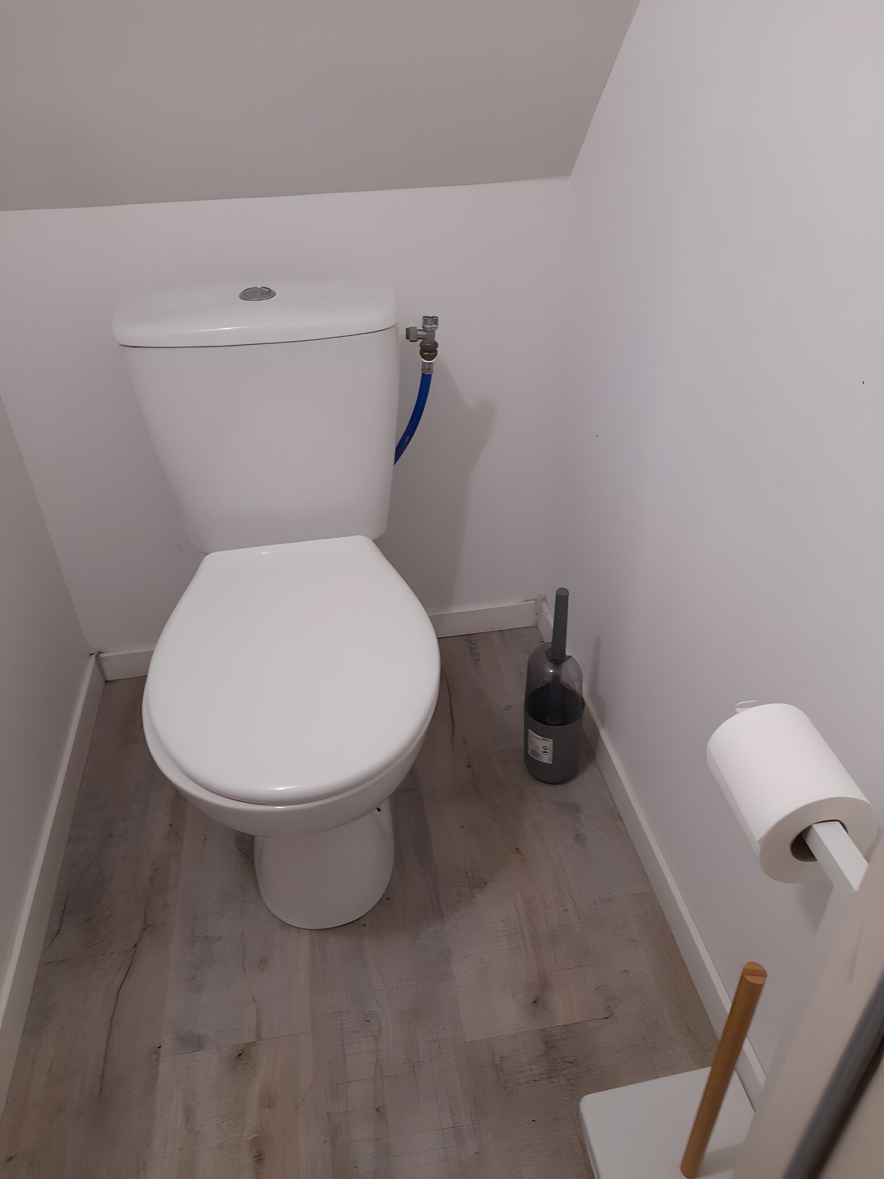 De petites toilettes blanches, avec un tuyau bleu raccordé à la cuvette, un porte-rouleau de papier toilette et un drain au sol.