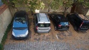 A parking area with five cars parked in a row, a cobblestone ground, and a brick wall.