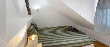 A small, sloped ceiling room with a bed, pillows, a bedside table, and a lamp.