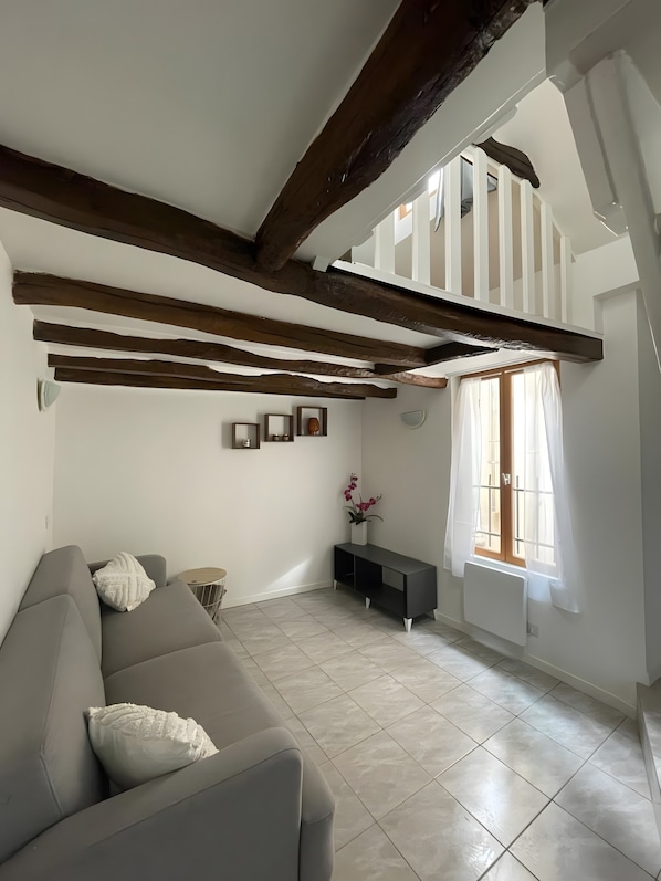A living room with a grey sofa, wooden beams, a white radiator, and a window with curtains.