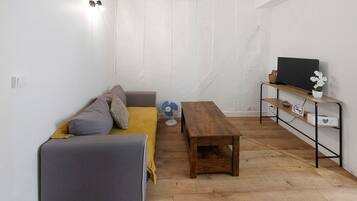 Une petite pièce agréable, avec un canapé gris, une table en bois et un téléviseur posé sur une étagère.